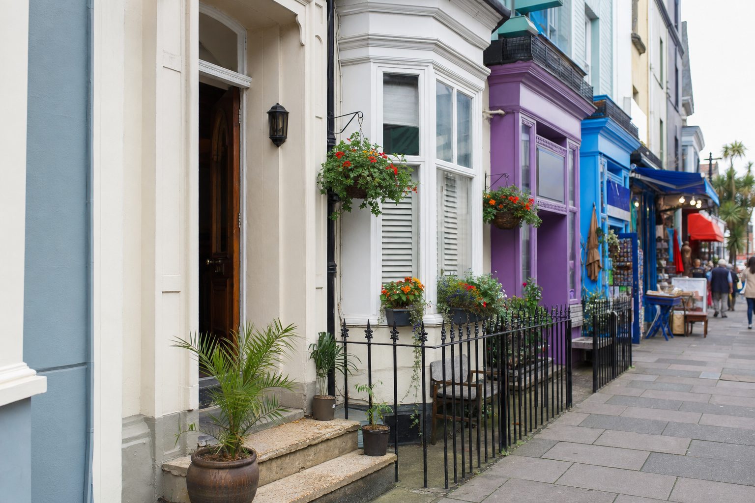 A row of pastel-painted terraced shopfronts with hanging flower baskets and iron railings on a quiet English high street