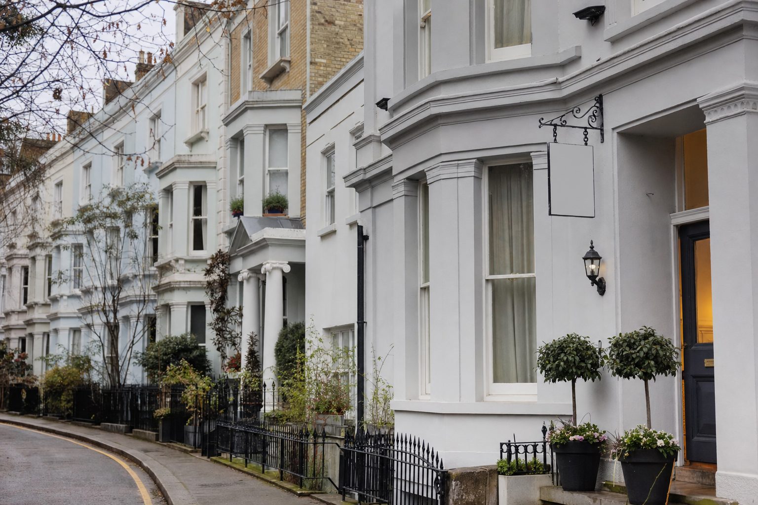 A row of pale Victorian terraced townhouses with bay windows, black iron railings, and potted topiary either side of a lit doorway