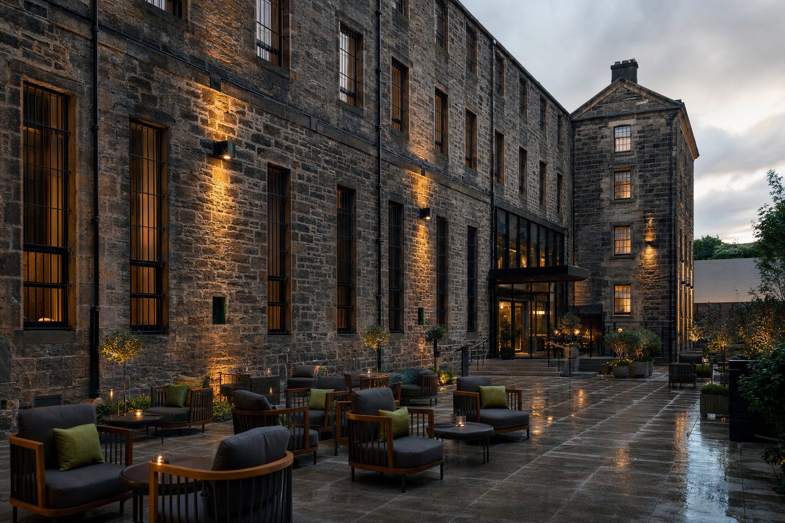 A converted stone building's courtyard at dusk, uplit walls and rain-slicked flagstones surrounding outdoor lounge seating with cushions