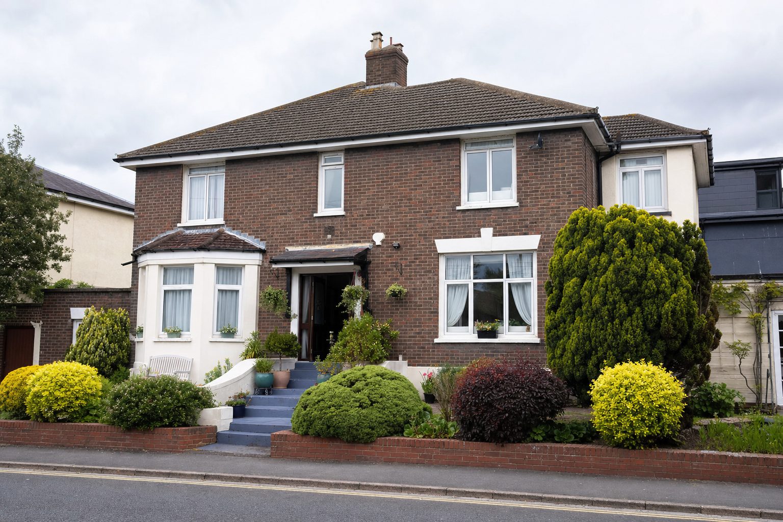 A two-storey brick house with a manicured front garden, clipped shrubs, and painted blue steps leading to the entrance
