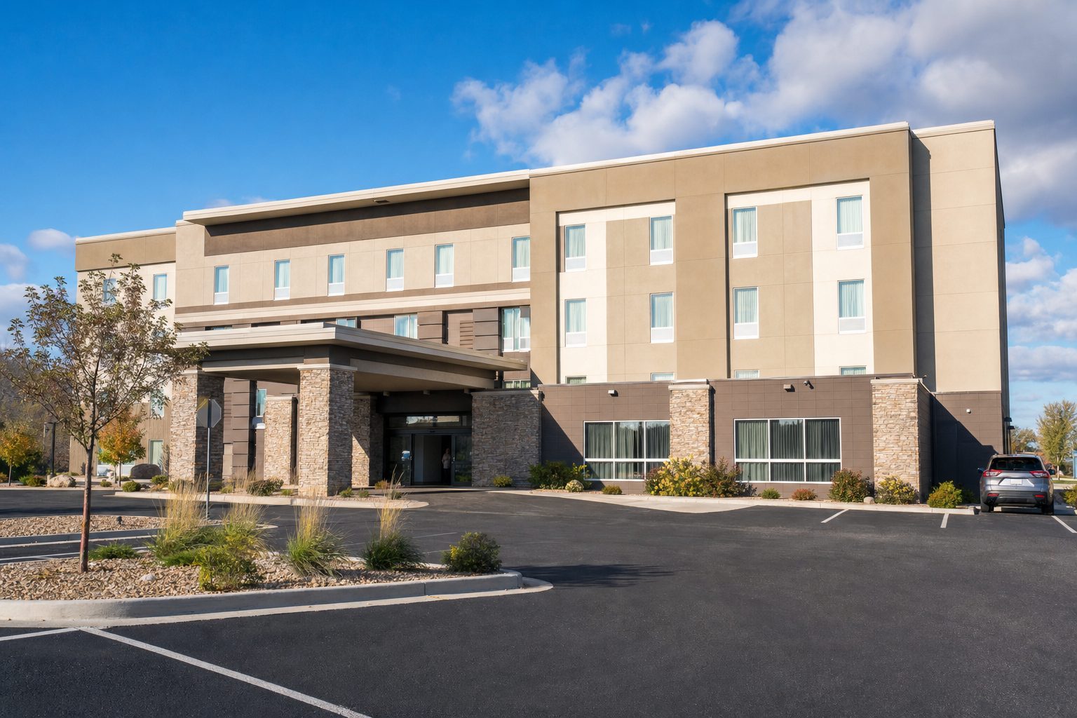 A modern low-rise budget hotel with a stone-pillared entrance canopy and a spacious car park under a bright blue sky